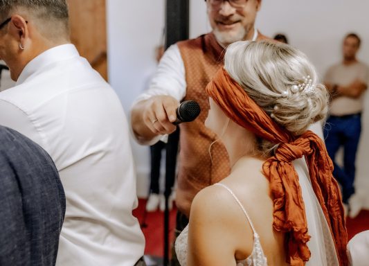 Mick moderiert ein Spiel, die Braut hat verbundene Augen und muss sagen was sie gerade tut. 